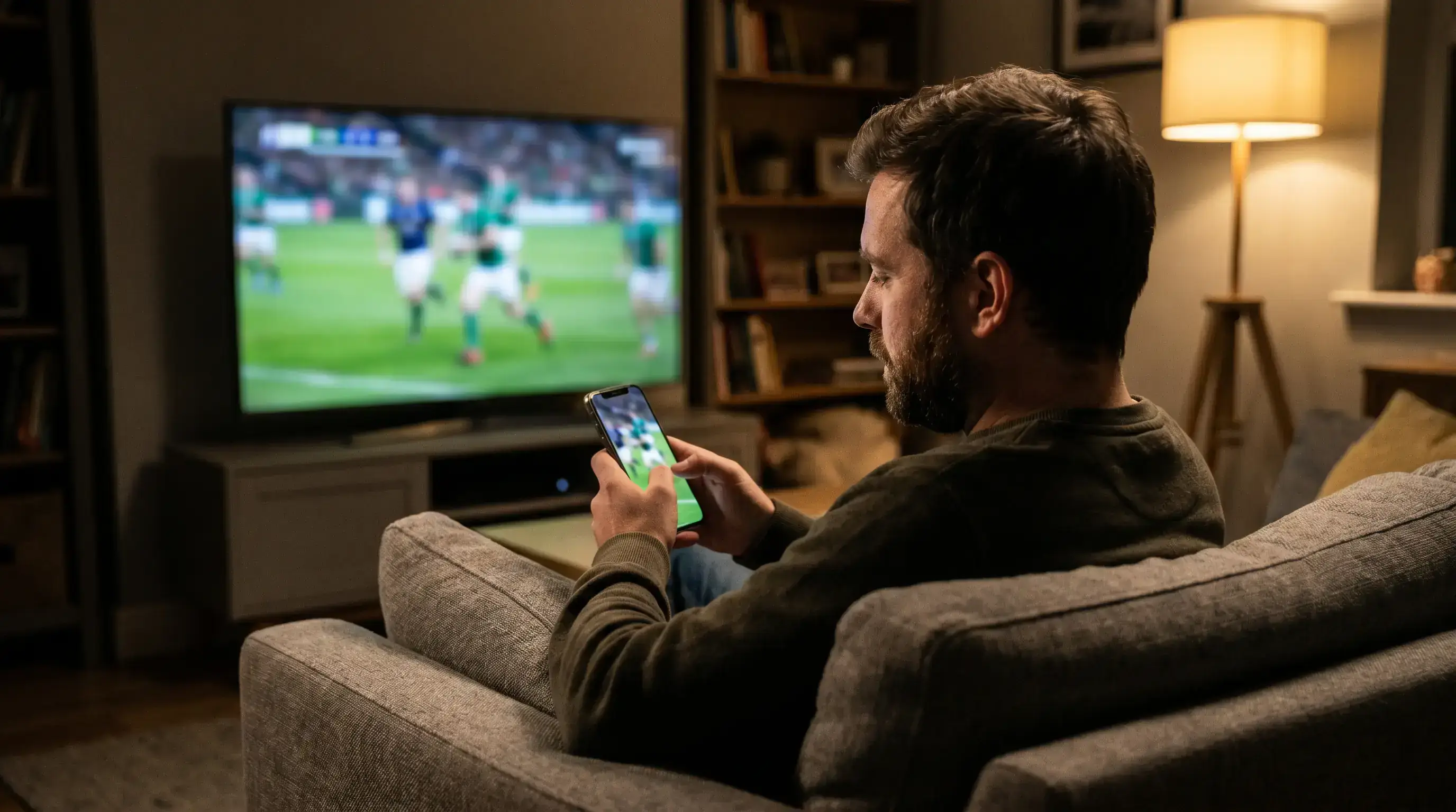 Person holding a smartphone showing live rugby match action while sitting on a sofa at home